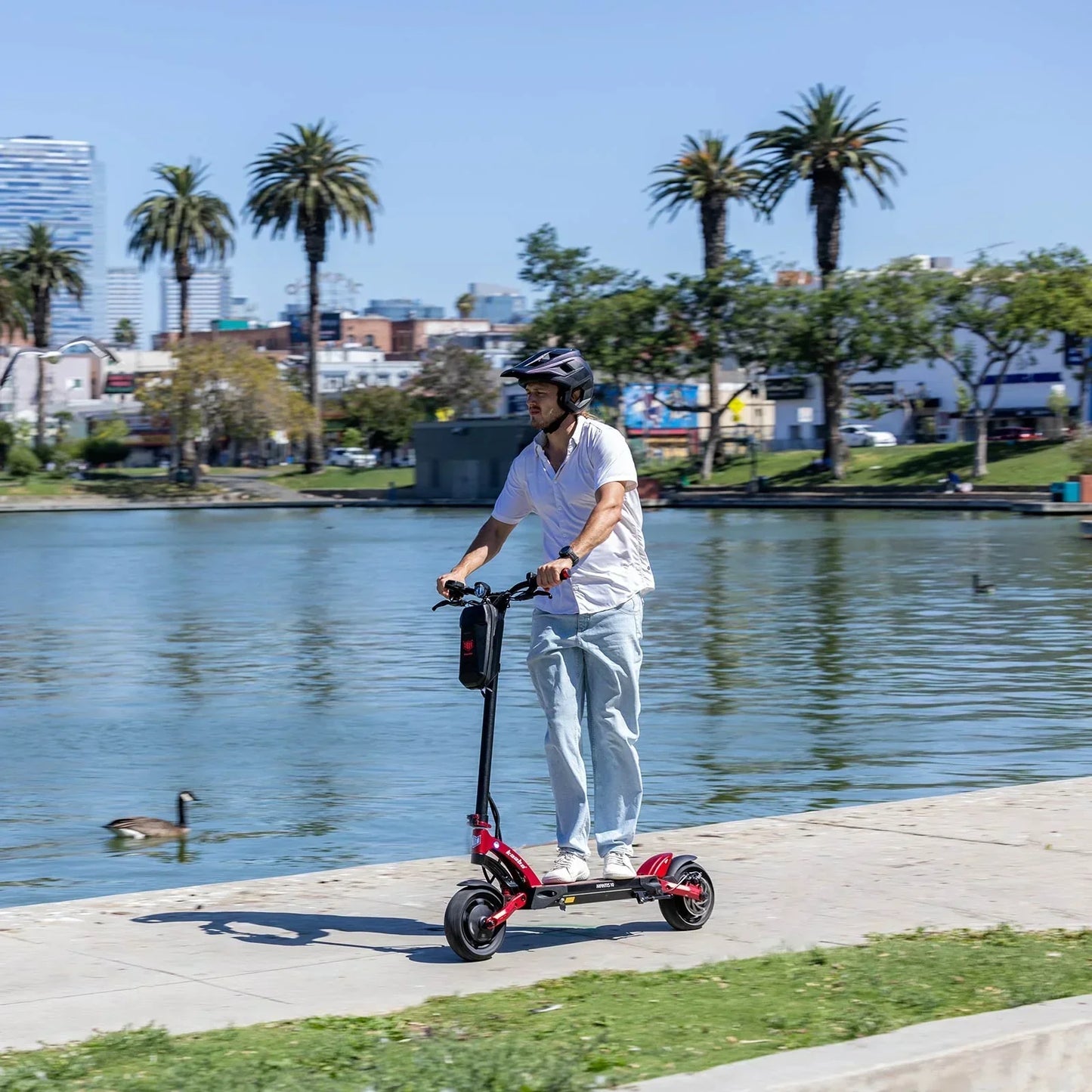 Person riding a red electric scooter in an urban setting with trees and buildings in the background.