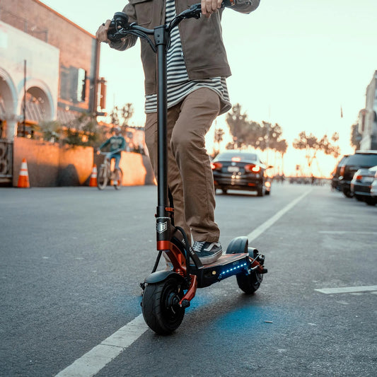 Person riding an electric scooter on a city street with buildings and cars in the background.