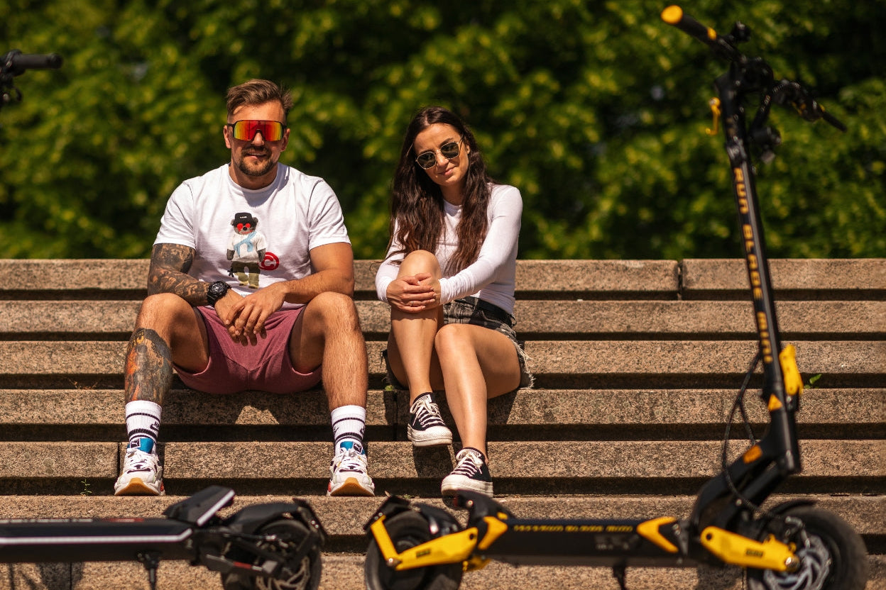Two people sitting on a bench with electric scooters beside them, surrounded by greenery.