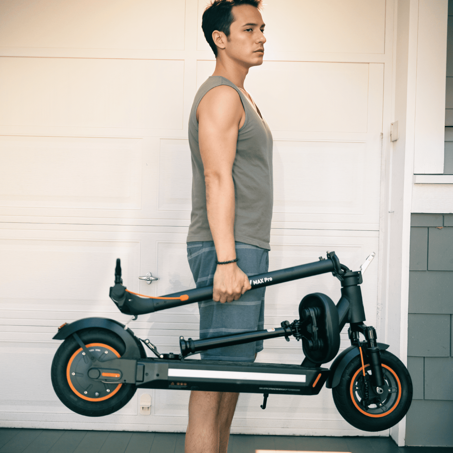 Man holding a black electric scooter with orange accents against a white wall.