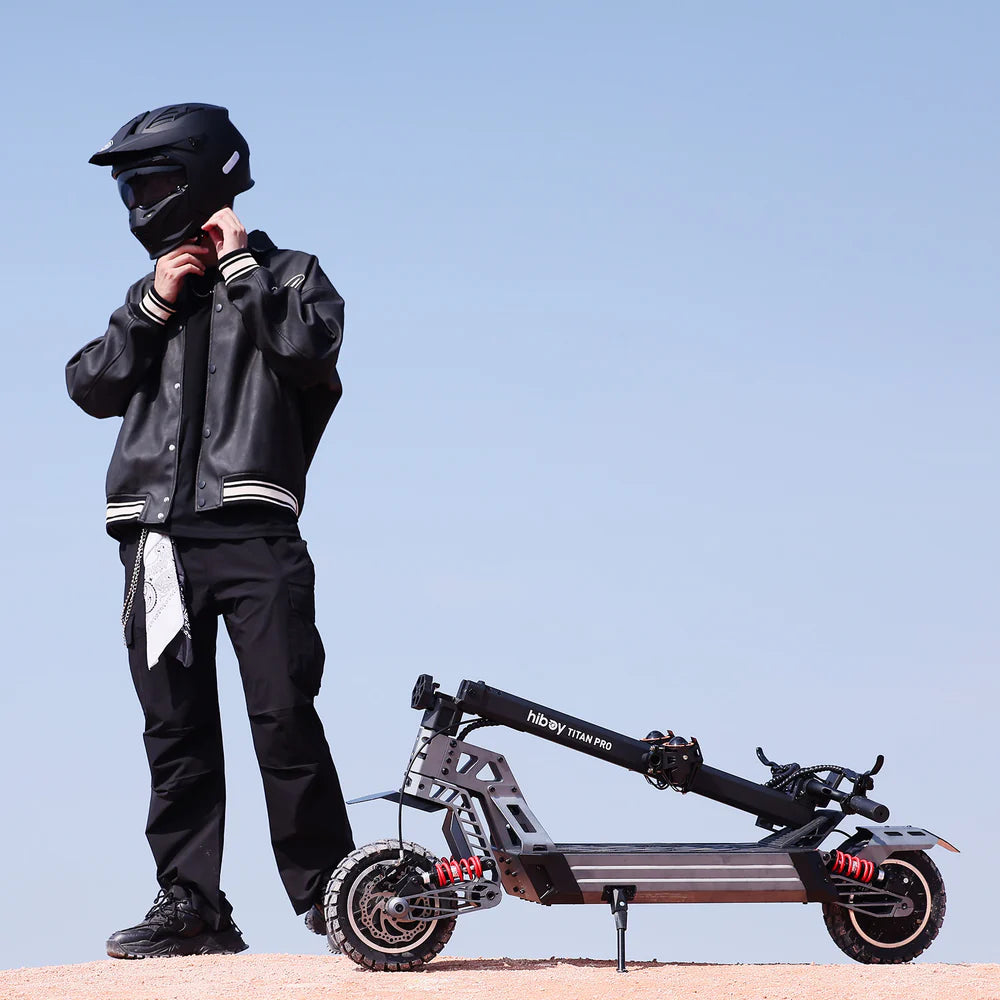 Person in black motorcycle gear standing next to a black electric scooter with a clear blue sky background