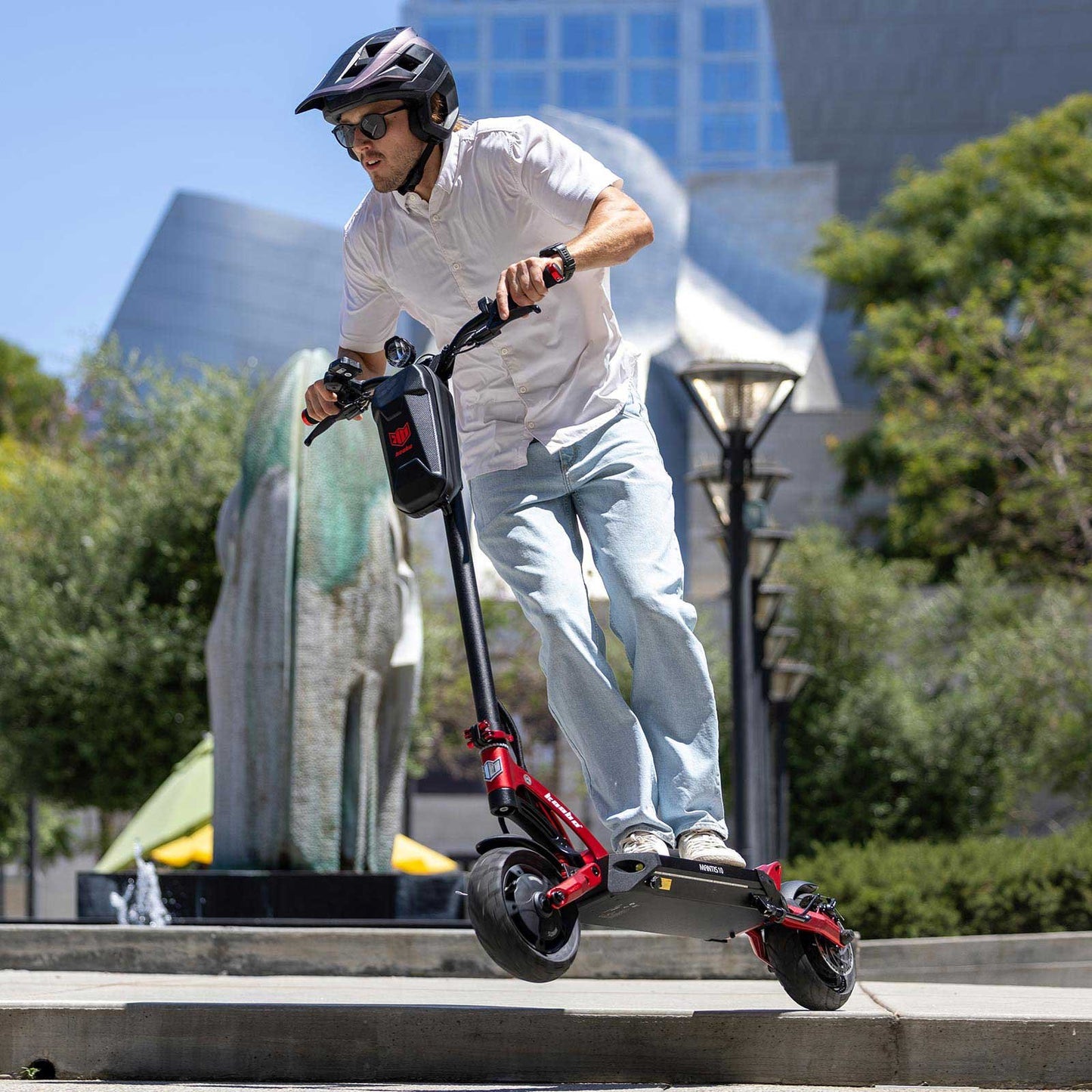 Person riding a red electric scooter in an urban setting with trees and buildings in the background.