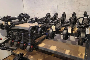 Multiple scooters with visible tags on a wooden platform against a white brick wall.